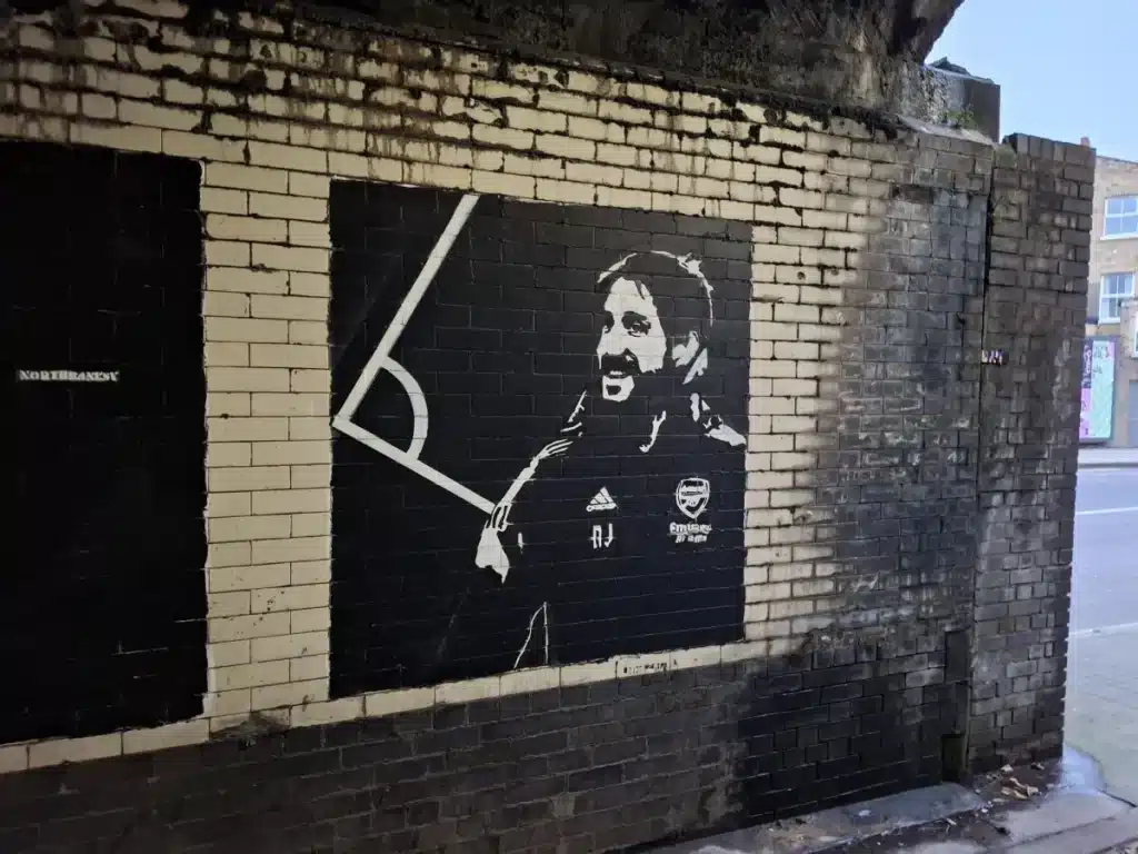 Stencil mural of a man in a jacket on a black square wall panel, on a brick urban alley.