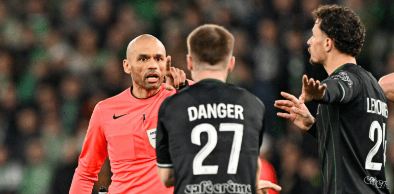 Referee in pink jersey arguing with two football players in black jerseys on a pitch, one with 'DANGER 27' on their back.