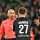 Referee in pink jersey arguing with two football players in black jerseys on a pitch, one with 'DANGER 27' on their back.