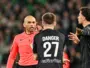 Referee in pink jersey arguing with two football players in black jerseys on a pitch, one with 'DANGER 27' on their back.