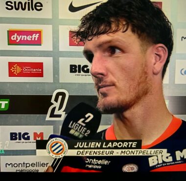 Football player Julien Laporte being interviewed after a match, holding a microphone in front of sponsor boards