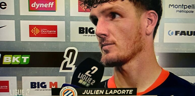 Football player Julien Laporte being interviewed after a match, holding a microphone in front of sponsor boards