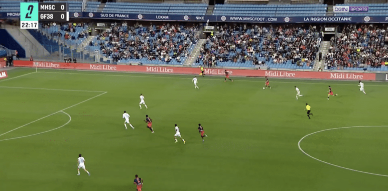 Soccer match in progress at a stadium; white and red teams vie for the ball near the right flank as the crowd watches, scoreboard shows MHSC 0-0 GF38 and 22:17.