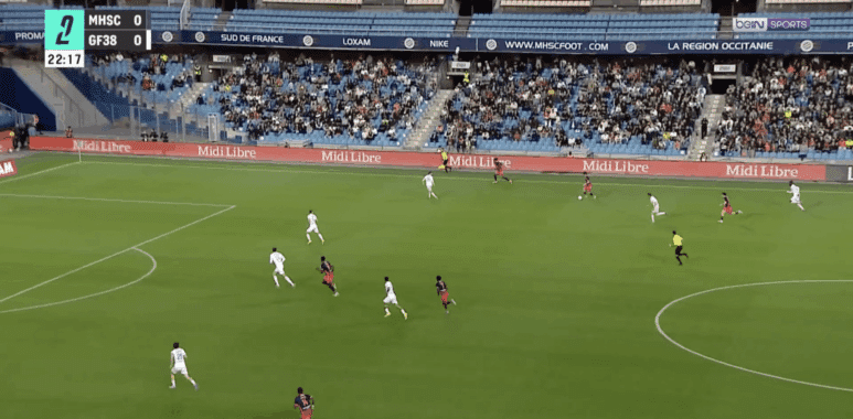 Soccer match in progress at a stadium; white and red teams vie for the ball near the right flank as the crowd watches, scoreboard shows MHSC 0-0 GF38 and 22:17.