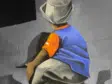 Back view of a person in a blue shirt and orange glove crouching against a gray wall, possibly painting graffiti on a mural.