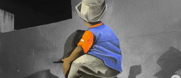 Back view of a person in a blue shirt and orange glove crouching against a gray wall, possibly painting graffiti on a mural.