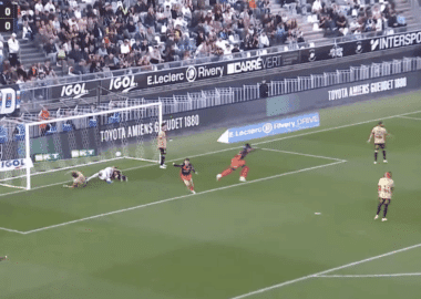 Soccer scene near goal: players in tan and red uniforms around the net as a ball sits by the goalkeeper on the ground, crowd in stands behind. BeIN SPORTS watermark visible.