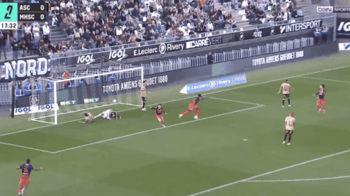 Soccer scene near goal: players in tan and red uniforms around the net as a ball sits by the goalkeeper on the ground, crowd in stands behind. BeIN SPORTS watermark visible.