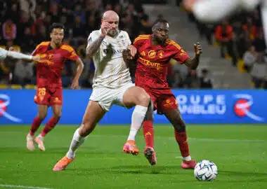 Two soccer players battle for the ball during a match, crowd watching in the stands behind them.