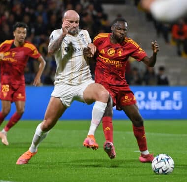 Two soccer players battle for the ball during a match, crowd watching in the stands behind them.