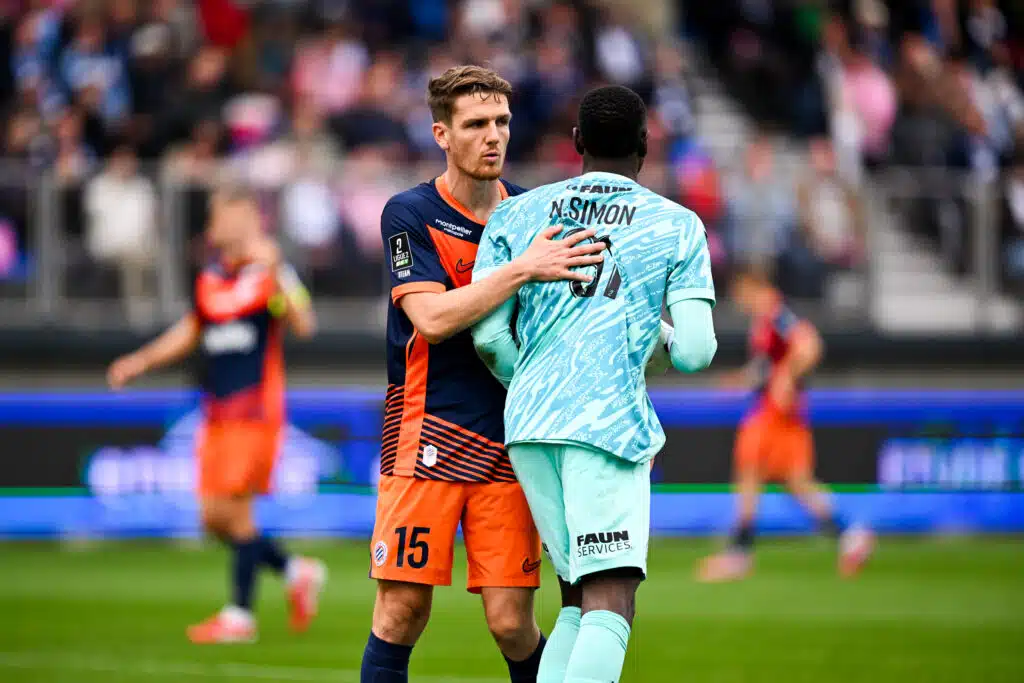 Two soccer players embrace on the field during a match, one in navy and orange and the other in light blue, with a blurred crowd in the background.