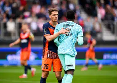 Two soccer players embrace on the field during a match, one in navy and orange and the other in light blue, with a blurred crowd in the background.