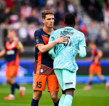 Two soccer players embrace on the field during a match, one in navy and orange and the other in light blue, with a blurred crowd in the background.