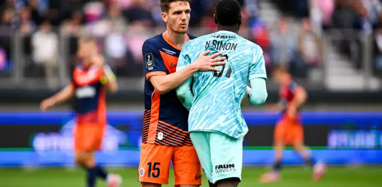 Two soccer players embrace on the field during a match, one in navy and orange and the other in light blue, with a blurred crowd in the background.