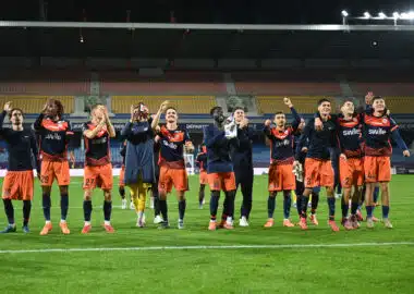 Soccer players in orange and navy celebrate on the field with arms raised and smiles, stadium stands behind them.