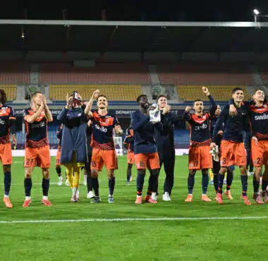 Soccer players in orange and navy celebrate on the field with arms raised and smiles, stadium stands behind them.
