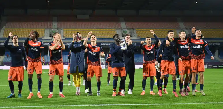 Soccer players in orange and navy celebrate on the field with arms raised and smiles, stadium stands behind them.