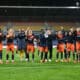 Soccer players in orange and navy celebrate on the field with arms raised and smiles, stadium stands behind them.