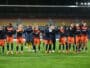 Soccer players in orange and navy celebrate on the field with arms raised and smiles, stadium stands behind them.