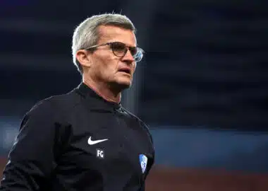 Man with gray hair and glasses in a black athletic jacket on a sports field, looking to the right.