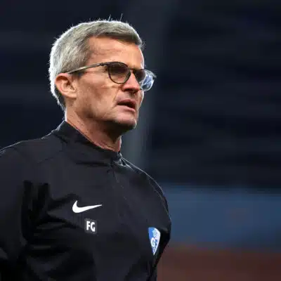 Man with gray hair and glasses in a black athletic jacket on a sports field, looking to the right.