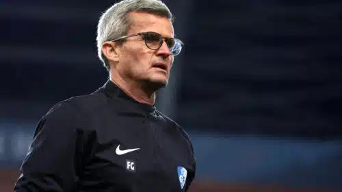 Man with gray hair and glasses in a black athletic jacket on a sports field, looking to the right.