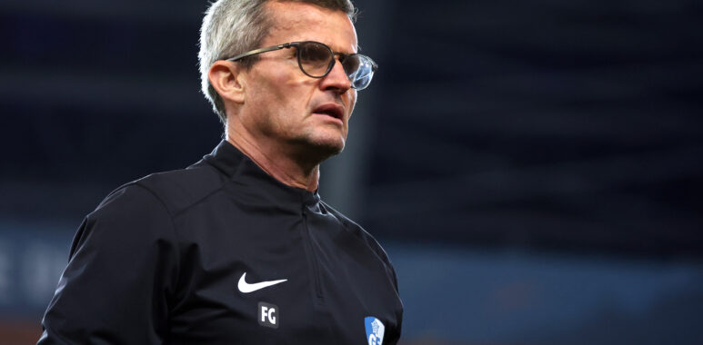 Man with gray hair and glasses in a black athletic jacket on a sports field, looking to the right.