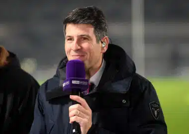 Man in a dark jacket holding a purple beIN SPORTS microphone and smiling at the camera.