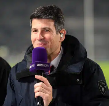 Man in a dark jacket holding a purple beIN SPORTS microphone and smiling at the camera.