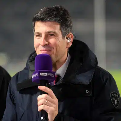 Man in a dark jacket holding a purple beIN SPORTS microphone and smiling at the camera.