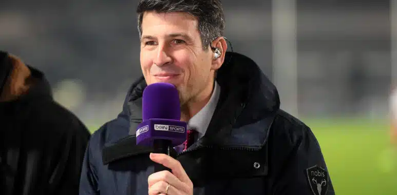 Man in a dark jacket holding a purple beIN SPORTS microphone and smiling at the camera.