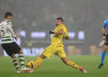 Soccer players in a rainy stadium duel for the ball, with a yellow-clad player about to kick it while a green-and-white defender blocks nearby.