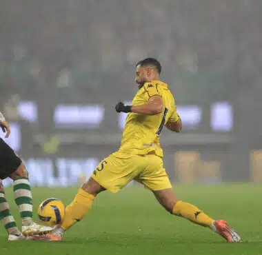 Soccer players in a rainy stadium duel for the ball, with a yellow-clad player about to kick it while a green-and-white defender blocks nearby.