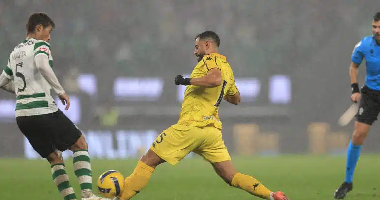Soccer players in a rainy stadium duel for the ball, with a yellow-clad player about to kick it while a green-and-white defender blocks nearby.