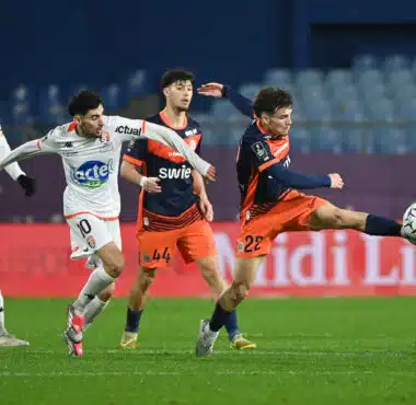 Four soccer players compete for the ball on a green pitch; white and orange kits, stadium seating in the background.