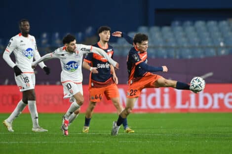 Four soccer players compete for the ball on a green pitch; white and orange kits, stadium seating in the background.
