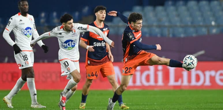 Four soccer players compete for the ball on a green pitch; white and orange kits, stadium seating in the background.
