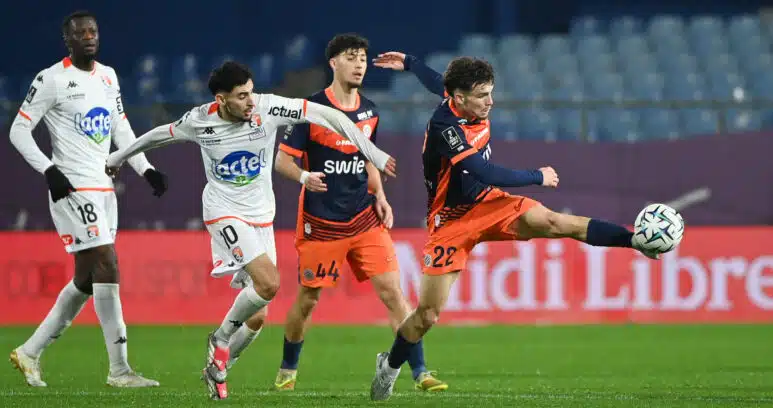Four soccer players compete for the ball on a green pitch; white and orange kits, stadium seating in the background.
