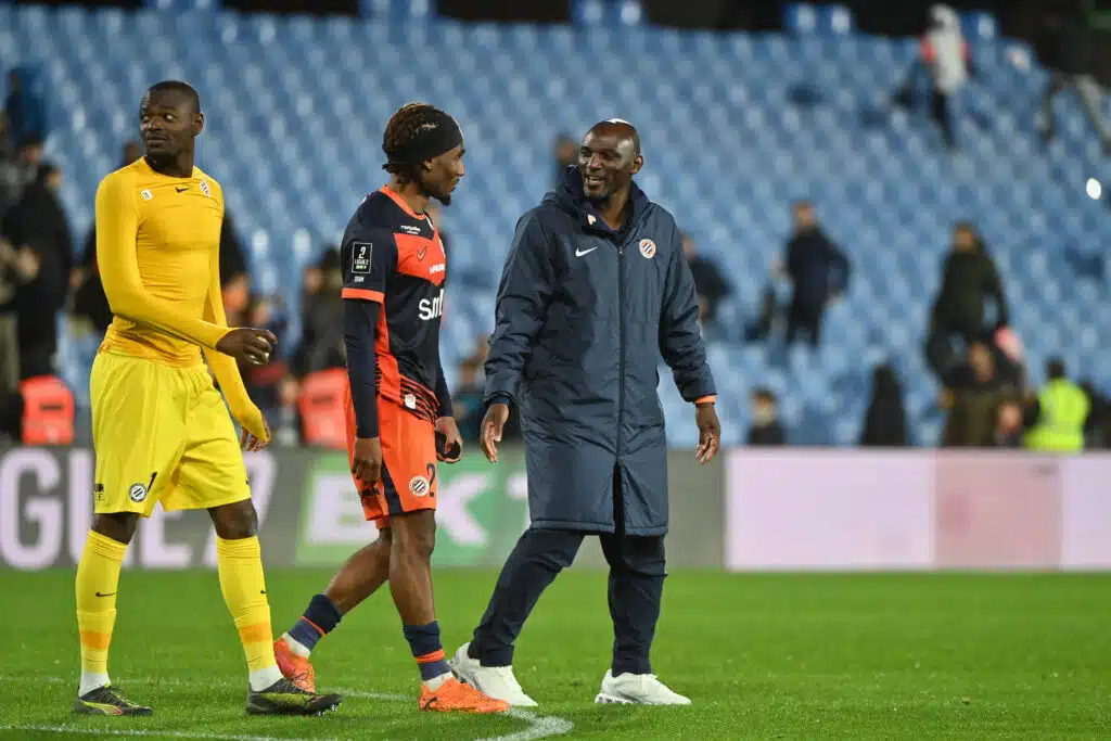 Football players and a coach walking on the pitch after a match, with blue stadium seats in the background.
