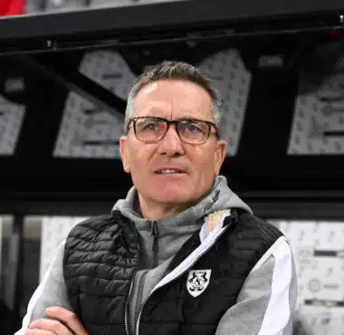 Man wearing glasses and a black puffer vest stands in front of a wall of boxes in a warehouse.**0