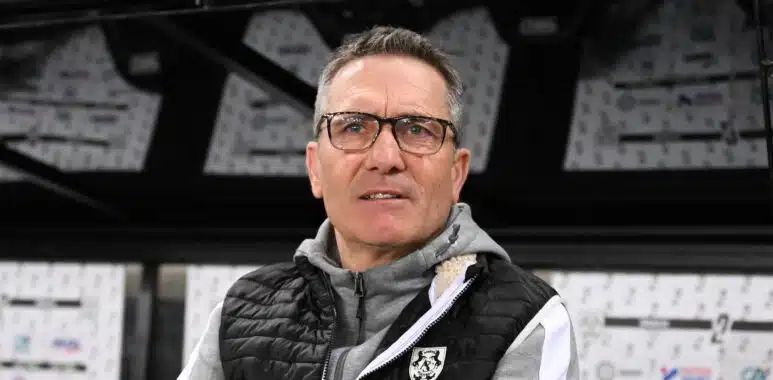 Man wearing glasses and a black puffer vest stands in front of a wall of boxes in a warehouse.**0