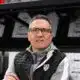 Man wearing glasses and a black puffer vest stands in front of a wall of boxes in a warehouse.**0