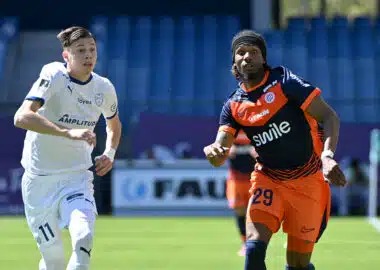 Two soccer players sprint side by side on a green field, one in white kit on the left and one in orange and navy on the right, chasing the ball.