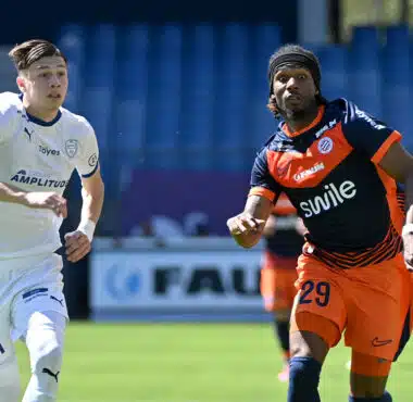 Two soccer players sprint side by side on a green field, one in white kit on the left and one in orange and navy on the right, chasing the ball.