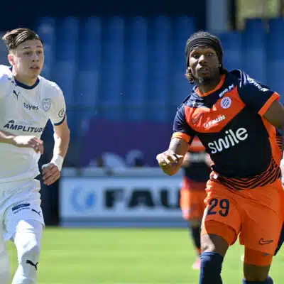 Two soccer players sprint side by side on a green field, one in white kit on the left and one in orange and navy on the right, chasing the ball.