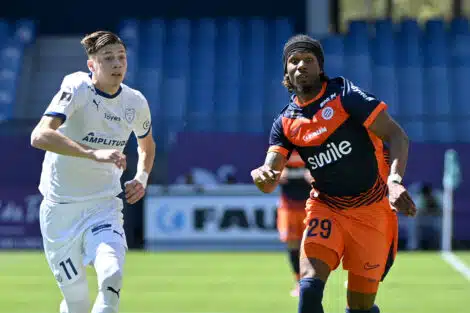 Two soccer players sprint side by side on a green field, one in white kit on the left and one in orange and navy on the right, chasing the ball.