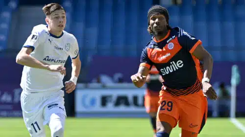 Two soccer players sprint side by side on a green field, one in white kit on the left and one in orange and navy on the right, chasing the ball.