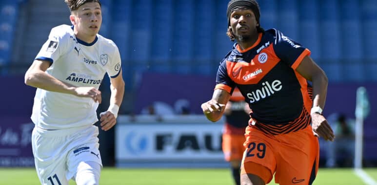Two soccer players sprint side by side on a green field, one in white kit on the left and one in orange and navy on the right, chasing the ball.