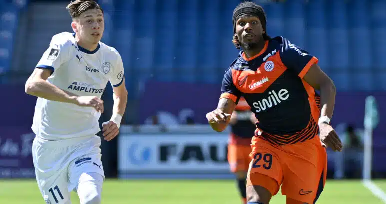 Two soccer players sprint side by side on a green field, one in white kit on the left and one in orange and navy on the right, chasing the ball.