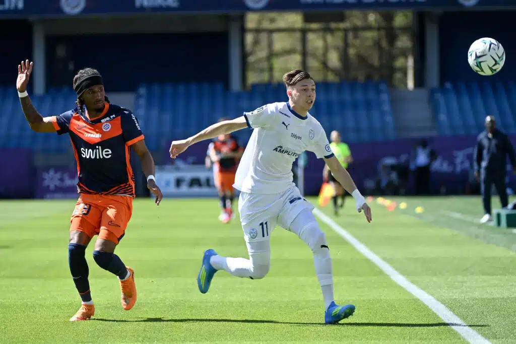 Two soccer players chase the ball during a daytime match on a green field, one in orange and navy and the other in white, with a ball mid‑air nearby